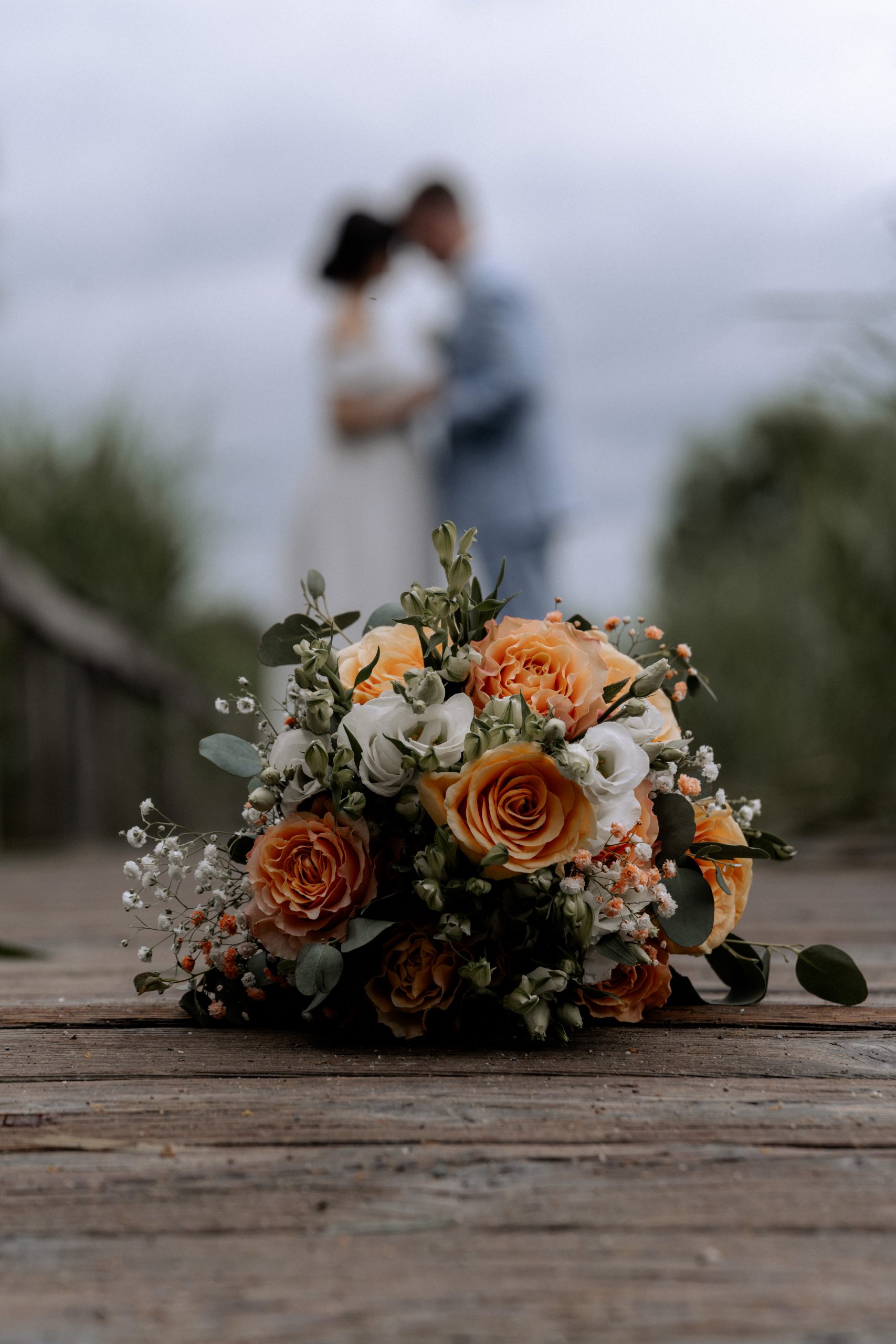 Hochzeitsstrauß mit Rosen und Schleierkraut vor unscharfem Brautpaar am See - Hochzeitsfotograf Pablo aus Thurgau